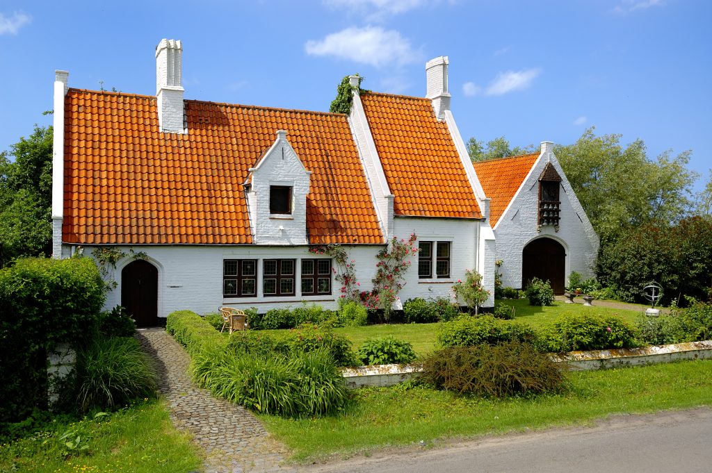 An image of a French provincial home. It is made of white painted stone, and has a steep, bright orange clay tile roof. There is a lack of symmetry and lots of ornamentation and vegetation.