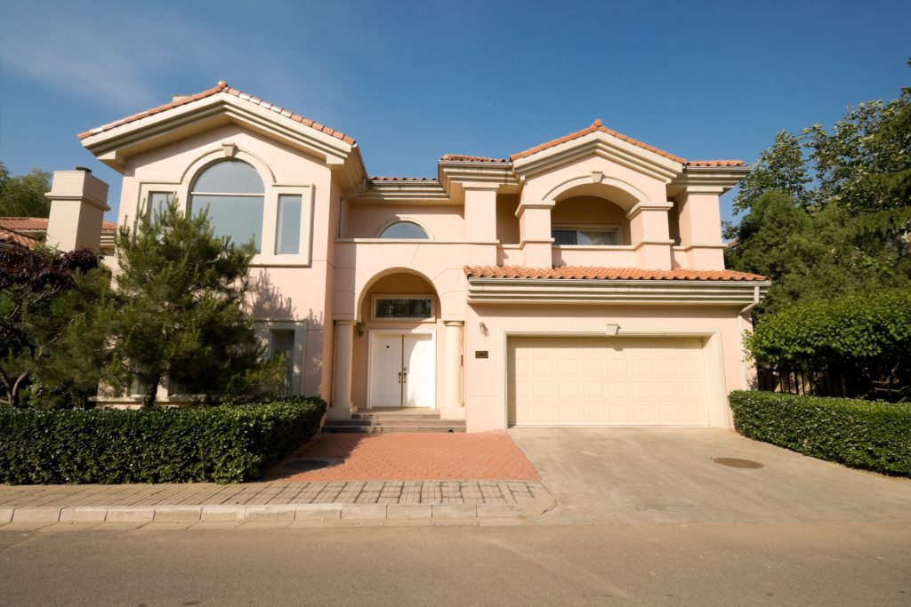 An image of a mediterranean style home. It is a salmon colored home, with large windows, a covered second-story balcony, and clay tiled roof. The attached garage is in the front, on the right side. 