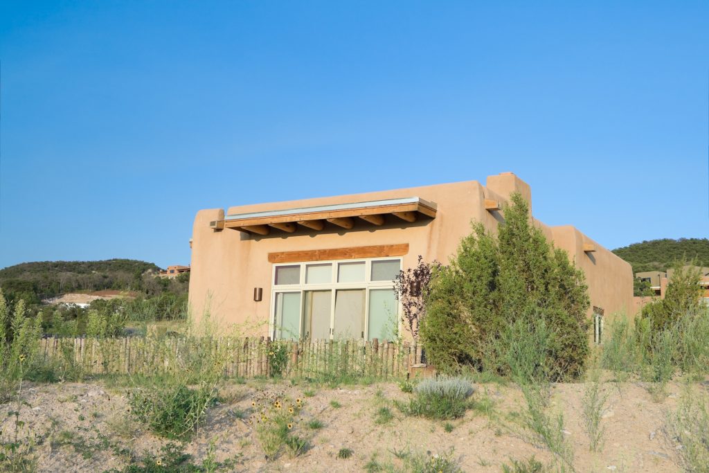 An image of a pueblo style home. This single family home is one tall story, made out of a light clay colored material. There is a block of windows out of the side of the house we see. It looks like a back yard, as there is a low fence made out of wooden posts. It is set in a desert, arid landscape somewhere. 